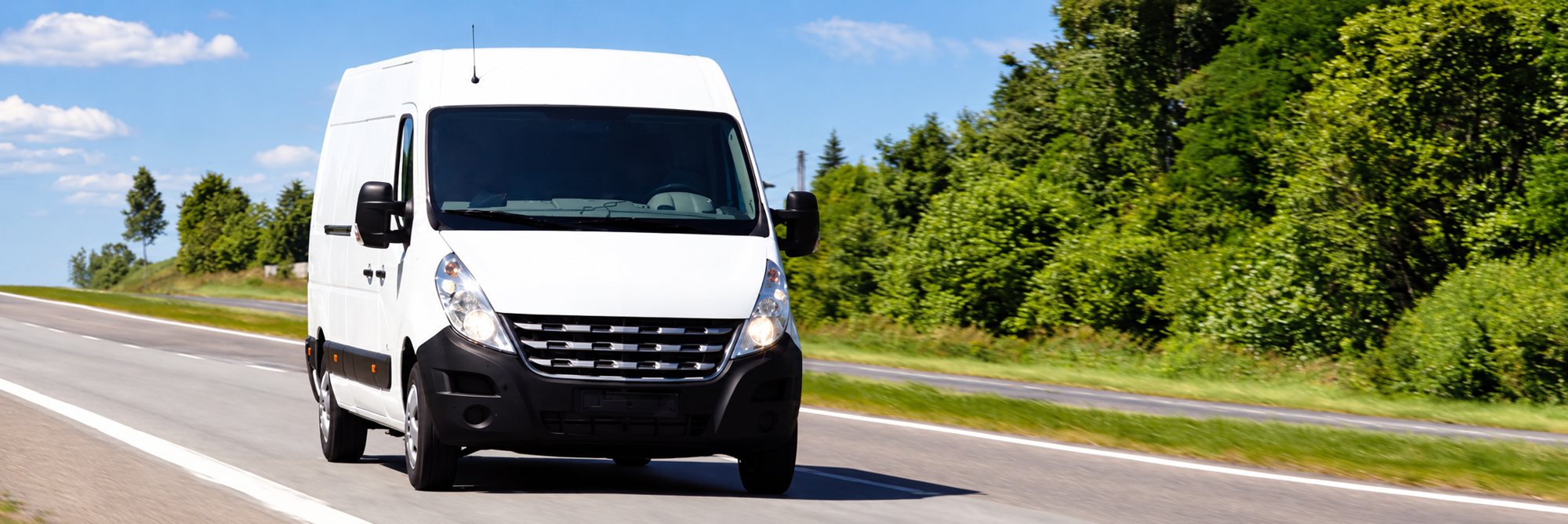Une camionnette blanche roule sur une route de campagne ensoleillée, bordée d’arbres verts.